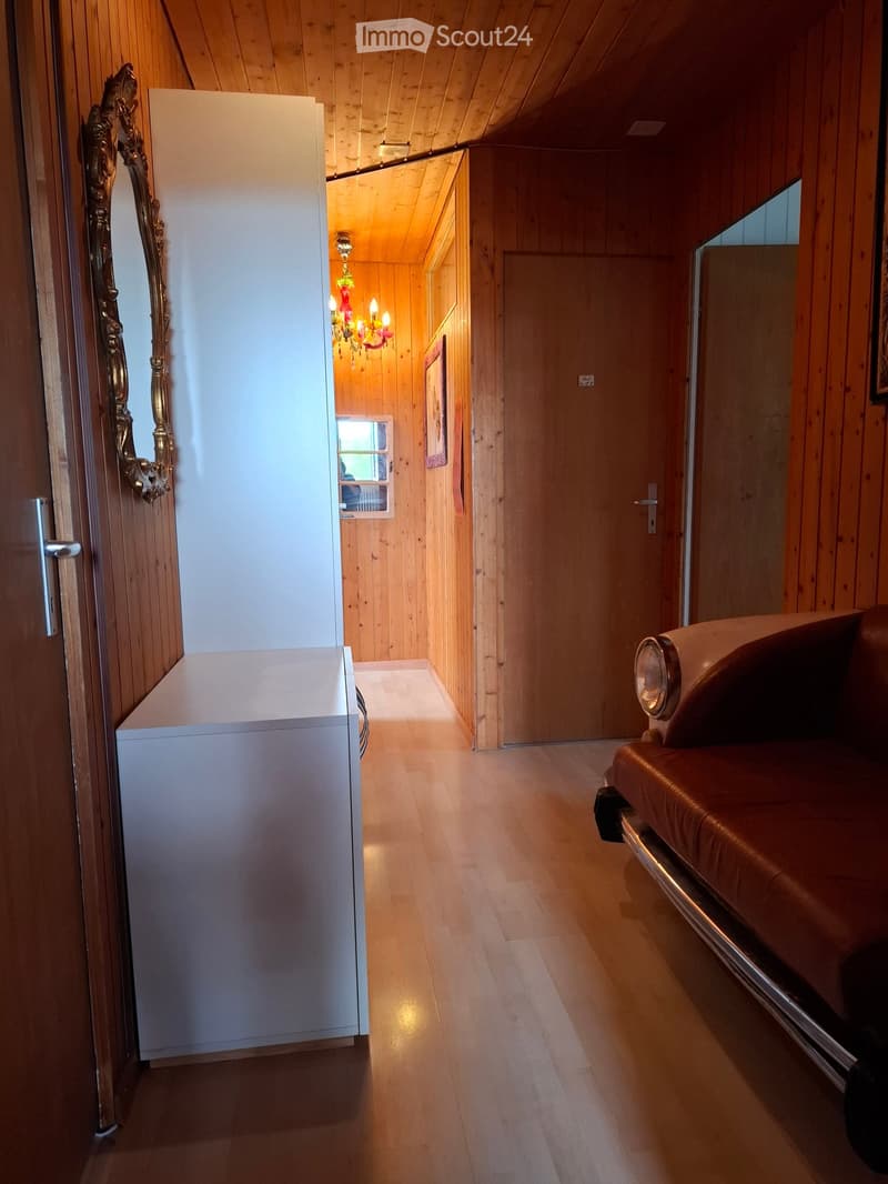 Wooden hallway with slanted ceiling, white cabinet, ornate mirror, chandelier, and a glimpse of a red leather sofa in the adjacent room.