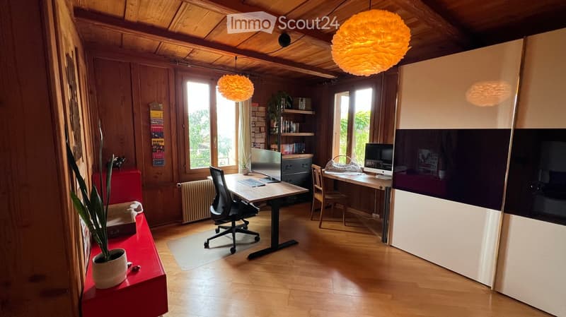 A modern, well-lit room with wooden floors and exposed wooden beams. It features two desks with chairs, multiple screens, a potted plant, and a shelf with books and decorative items.