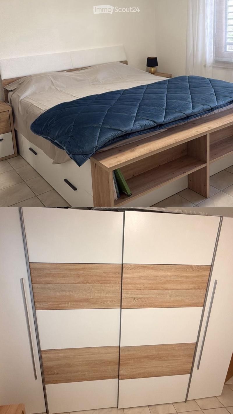 Modern bedroom with a wooden bed frame, built-in storage, and a nightstand with a lamp. The bed is neatly made with a blue duvet and white sheets.