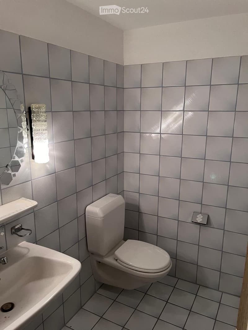 A small bathroom with grey tiled walls and floor, featuring a toilet, a sink with a mirror, and a faucet.