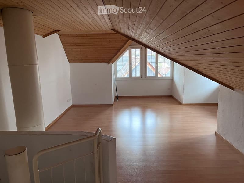 Empty room with wooden flooring and white walls, featuring a slanted wooden ceiling and large windows at the far end.