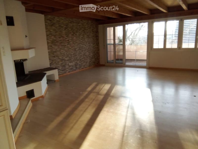 Empty room with wooden floor, exposed wooden ceiling beams, stone accent wall, large windows, and sliding glass doors leading to a balcony.
