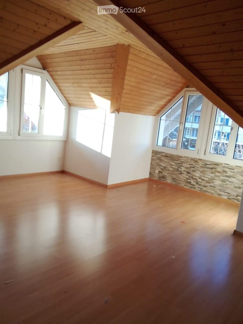 Empty attic room with wooden floors, white walls, and large windows, featuring exposed wooden beams on the ceiling.