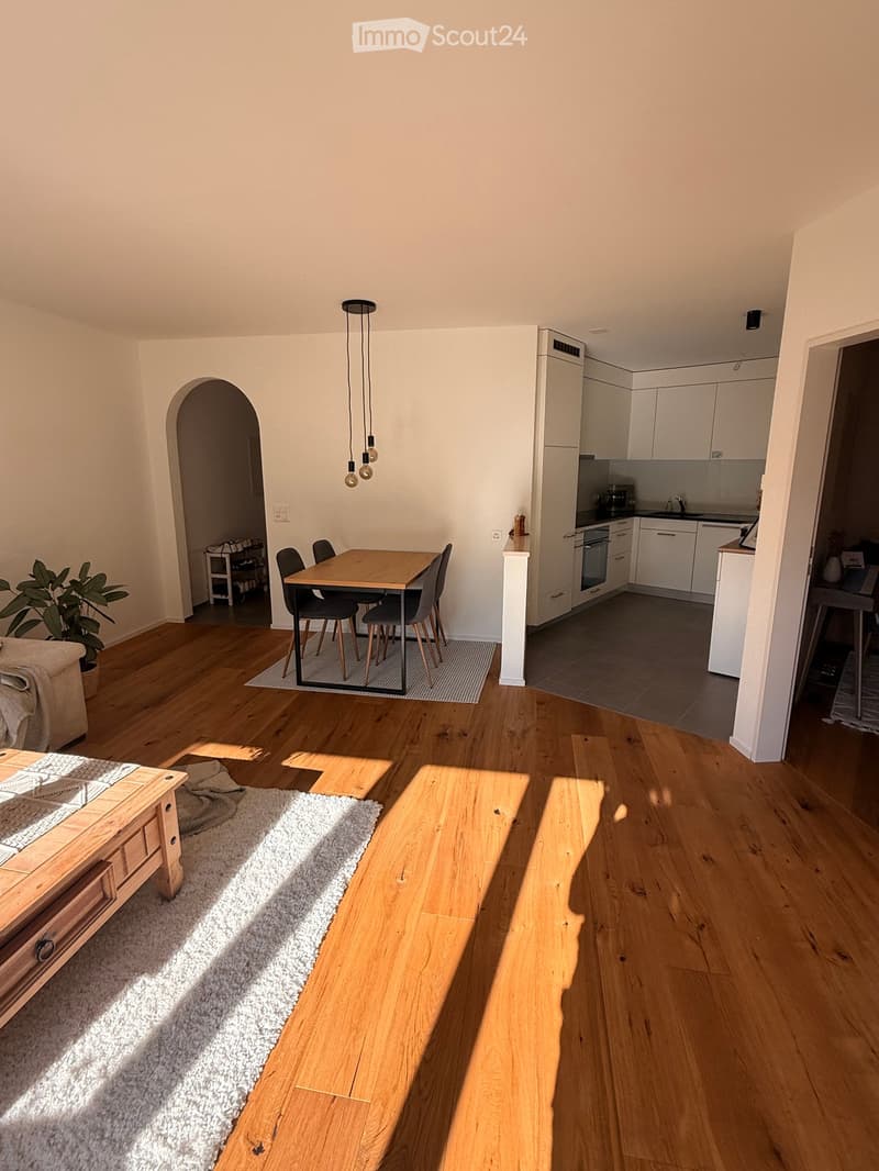 Bright living and dining area with wooden flooring, white walls, and modern kitchen in the background. Features a sofa, coffee table, dining table with chairs, and a bench.
