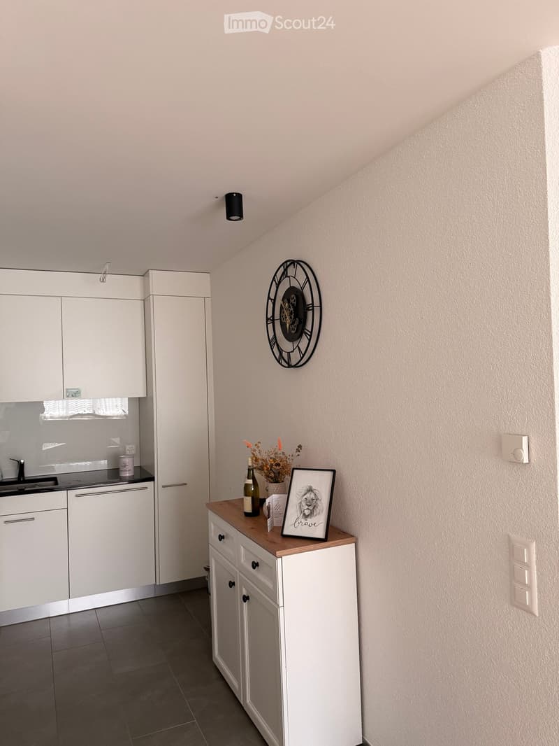 A small, modern kitchen area with white cabinets, a sink, a small table with a picture frame, a bottle, and a flower vase, and a clock on the wall.