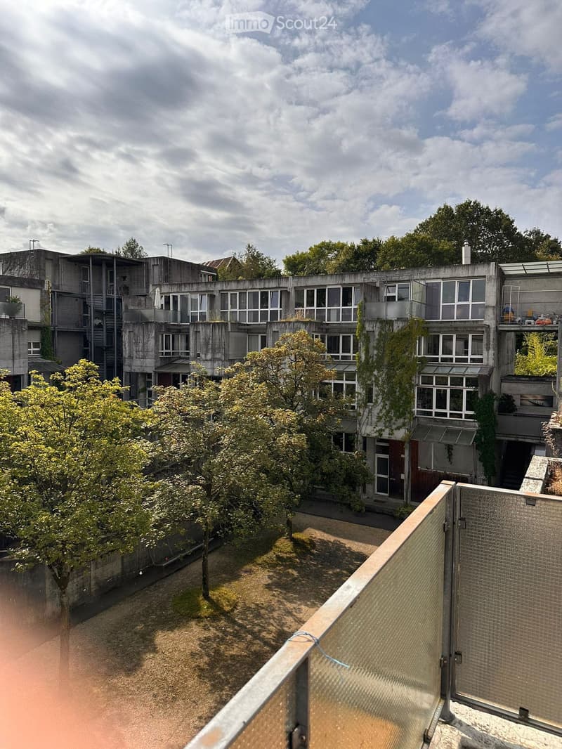 Modern multi-story building with large windows and balconies, surrounded by trees and greenery.