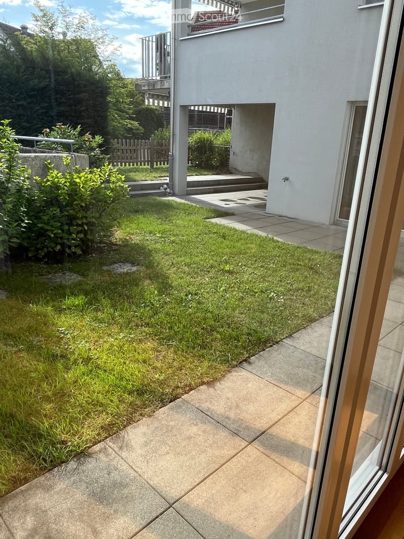 Garden area with grass, concrete patio, small steps leading to a house entrance, and surrounding greenery.