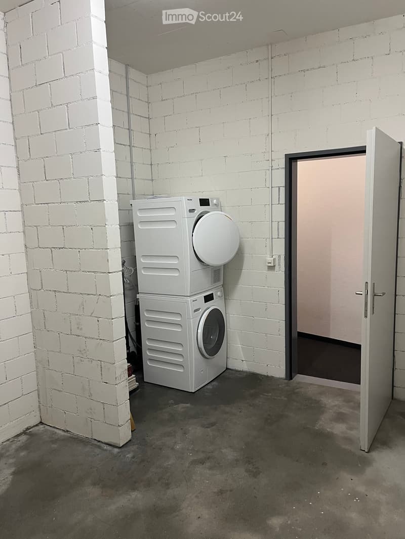 Empty room with white brick walls, concrete floor, and a door leading to another space. A white washing machine and dryer are stacked against the wall.