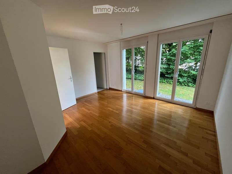 Empty room with wooden flooring, white walls, large sliding glass doors leading to a green outdoor area, and a visible doorway to another room.