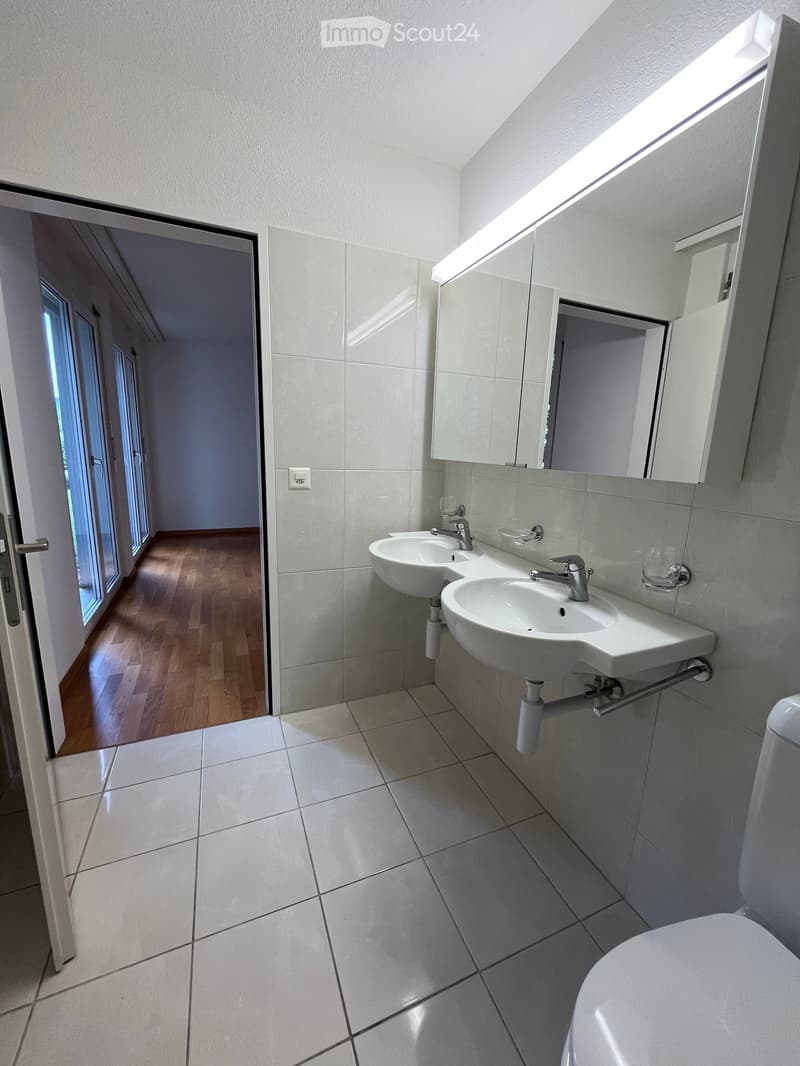 Modern bathroom with white tiled walls and floor, double sink vanity, large mirror, and a toilet. The room has a skylight window providing natural light.