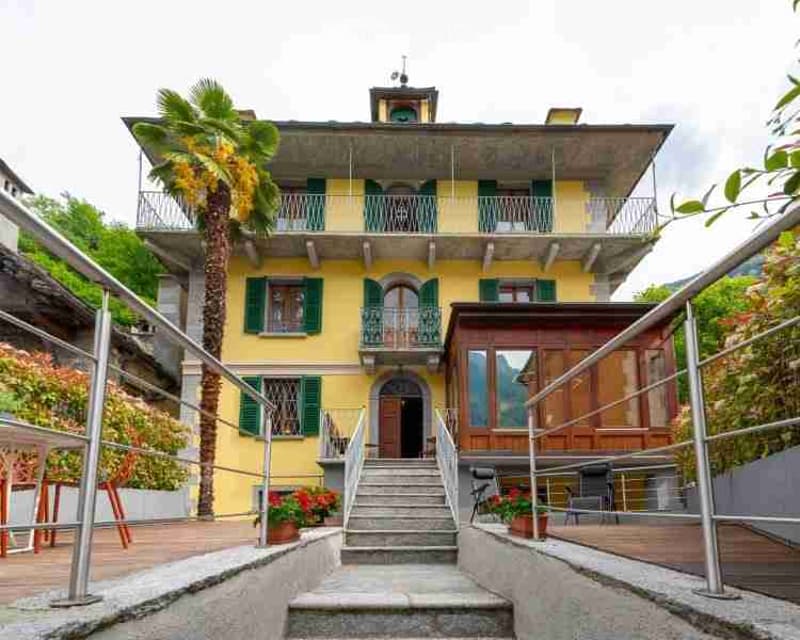 A two-story yellow building with green shutters and balconies, featuring a staircase leading to the entrance and a small wooden annex nearby.