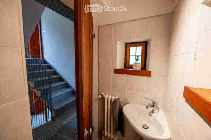 Small bathroom with a white sink, a radiator, and a towel hanging on it. A small window with a wooden frame is above the sink, and a wooden shelf is mounted on the wall to the right. An open wooden door leads to a staircase.