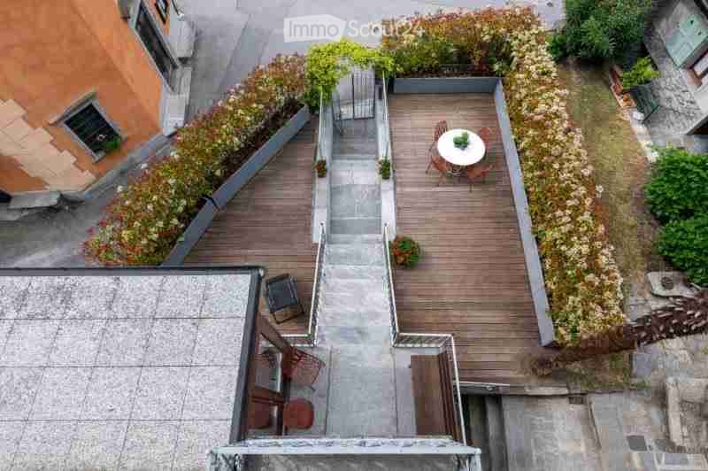 A small, well-maintained rooftop terrace with wooden decking, a round table with chairs, and a few potted plants. The space is bordered by low hedges and features a staircase leading up to the terrace.