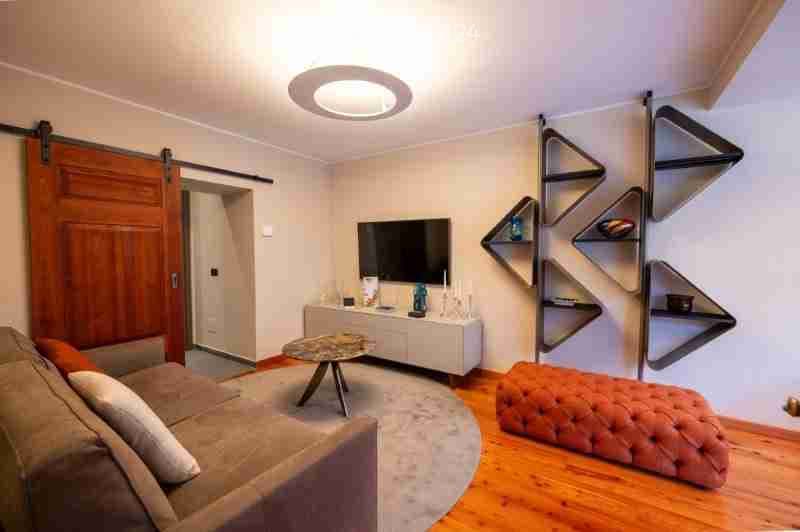 Modern living room with a grey sofa, ottoman, coffee table, TV stand, and wall-mounted shelves. The room features wooden flooring and a large round ceiling light.