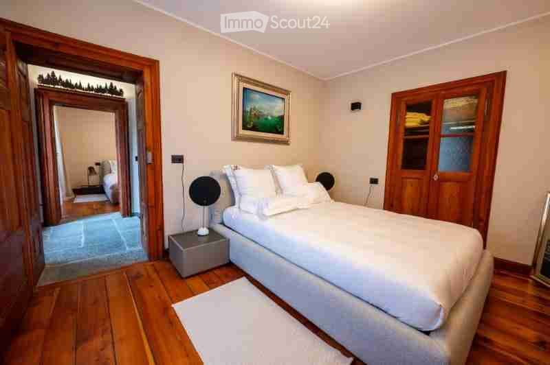 Cozy bedroom with wooden flooring, a large bed with white linens, a nightstand with a lamp, a picture frame on the wall, and a wooden wardrobe with glass panels.
