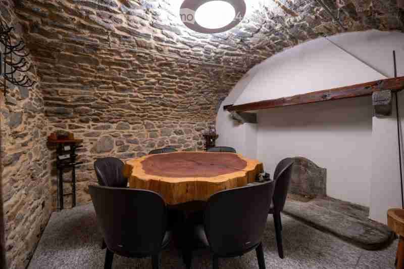 A rustic dining area with a round wooden table and six chairs, set against stone walls and a vaulted ceiling.