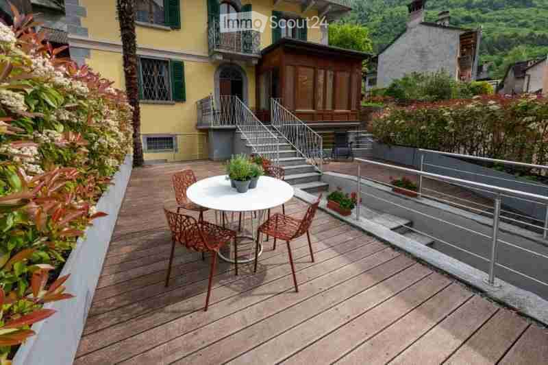 Wooden balcony with a small round table and four chairs, surrounded by greenery and a staircase leading to a building.