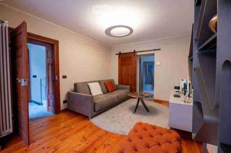 Modern living room with a grey sofa, orange and white cushions, a round coffee table, an orange ottoman, and a shelf with decorative items. The room features wooden flooring and large windows with wooden shutters.