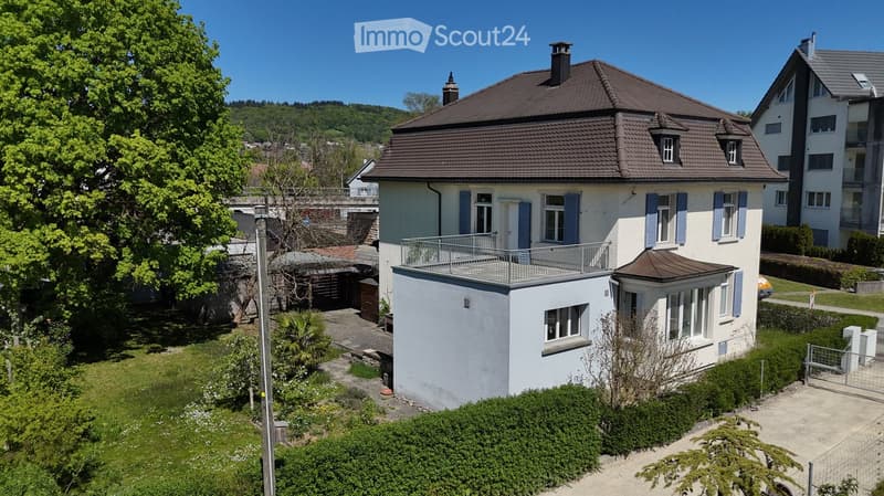 Two-story house with a brown tiled roof, white walls, blue shutters, and a balcony on the upper level. The house is surrounded by greenery and has a small garden area.