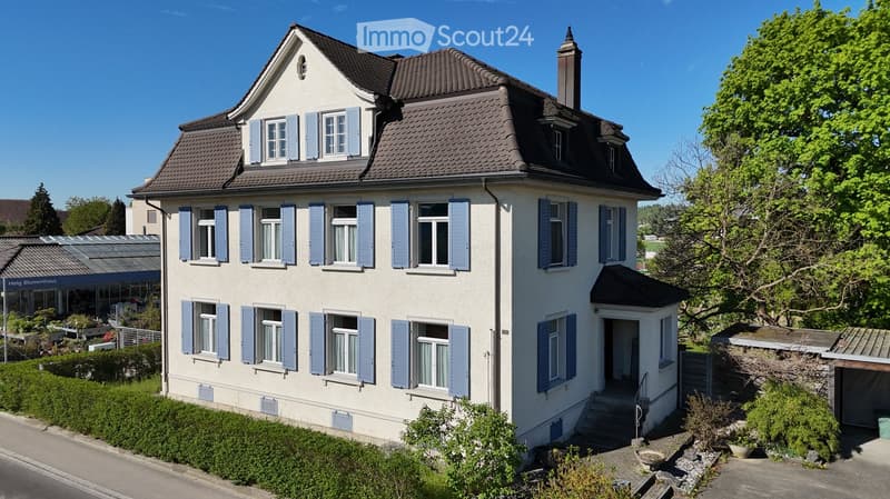 Two-story house with light-colored walls, multiple windows with shutters, and a chimney. The house has a well-maintained garden with greenery and a small entrance staircase.