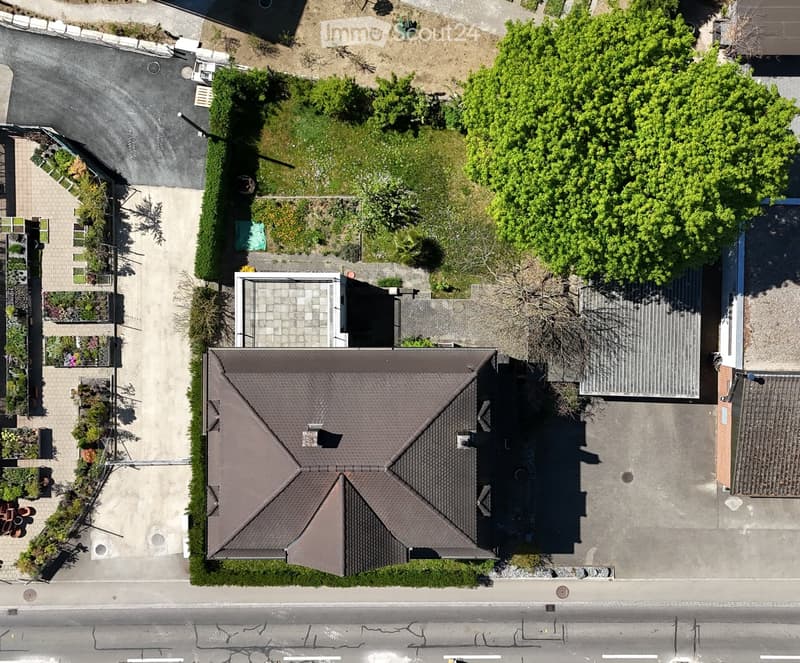 Aerial view of a two-story house with a dark brown shingled roof, surrounded by a well-maintained garden and driveway.