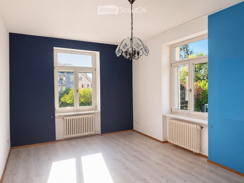 Empty room with wooden flooring, white walls, dark blue accent wall, large windows, radiator, and a decorative chandelier.