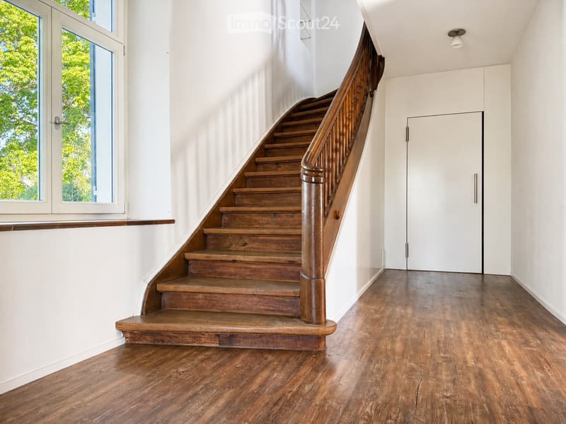Spacious hallway with wooden flooring, a wooden staircase with a polished handrail, and a white door leading to another room.