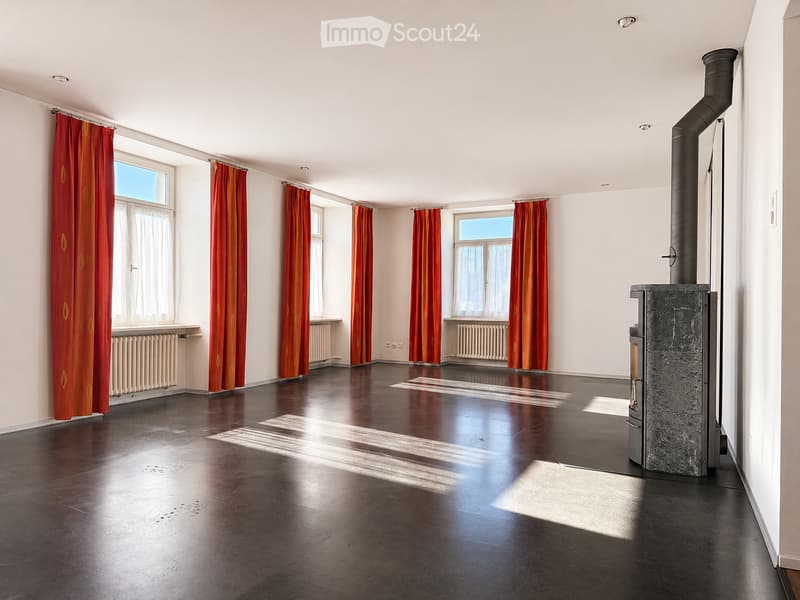 Empty room with dark wooden flooring, white walls, multiple windows with red curtains, and a stone fireplace with a chimney.