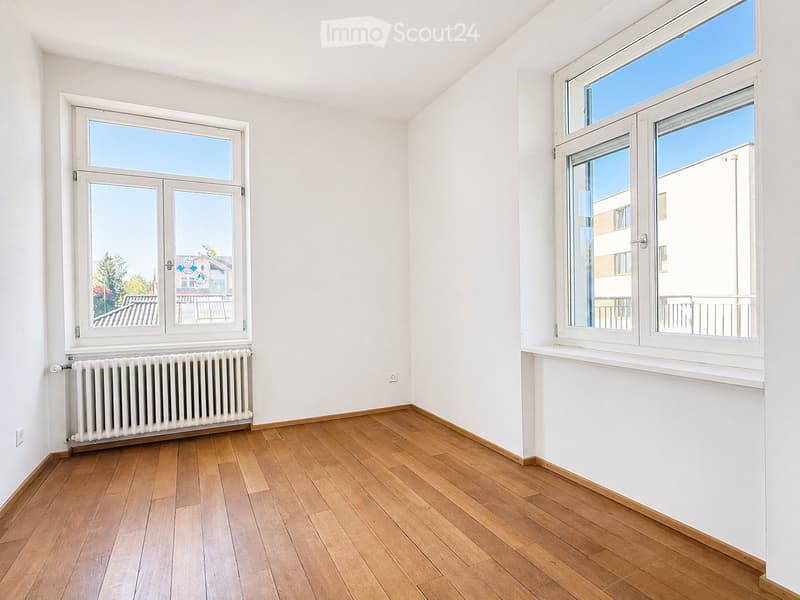 Empty room with wooden flooring, white walls, and large windows offering a view of the outside. A radiator is visible under one window.