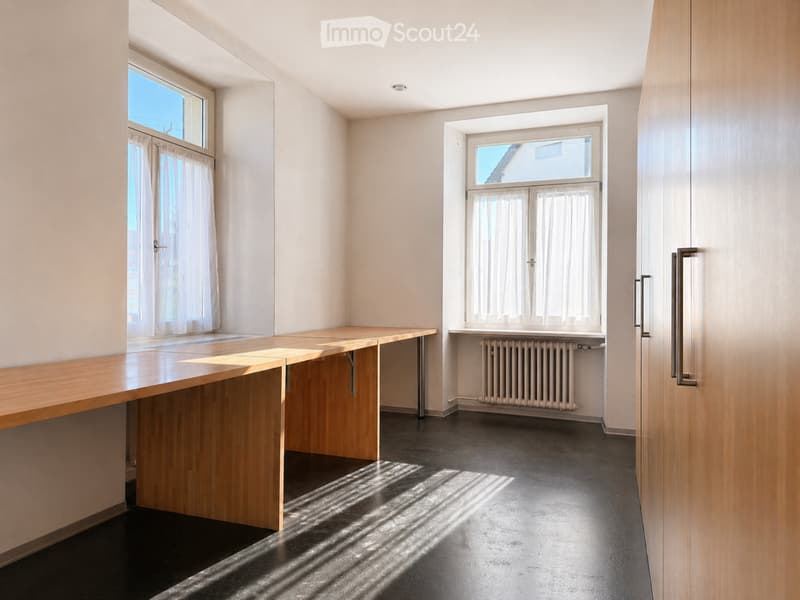 Empty room with light-colored walls, dark floor, large windows with white curtains, and a wooden bench against one wall.