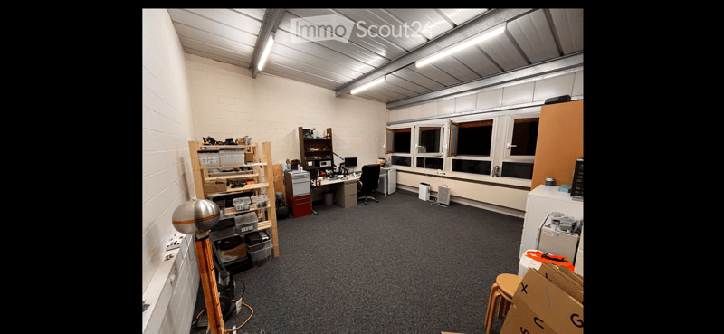 Office space with a desk, chair, shelves, and multiple stools. The room has white brick walls, large windows, and a gray carpeted floor.