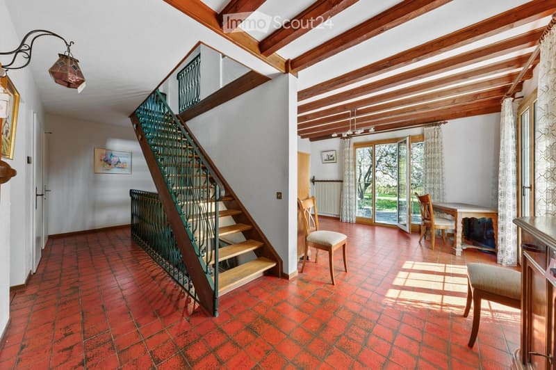Staircase with wooden steps and green wrought-iron railing, leading to an open living area with red tiled floor, wooden ceiling beams, and large windows with sheer curtains. Visible furniture includes chairs and a table.