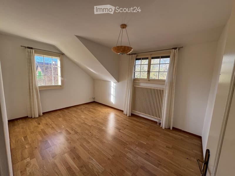 Empty attic room with wooden flooring, large windows, and a radiator. The room features a sloped ceiling and natural light from multiple windows.