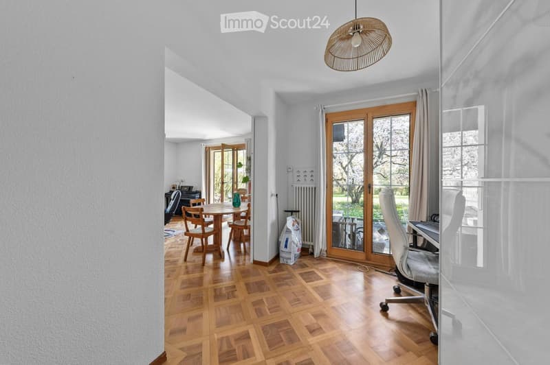 Bright, open-plan living and dining area with wooden flooring, large glass doors leading to a garden, a hanging woven pendant light, a white office chair, a laptop on a desk, and a round wooden dining table with four wooden chairs.