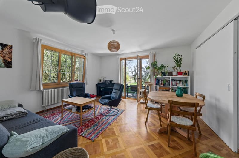 Bright living and dining area with wooden flooring, large windows, and sliding glass doors leading to a patio. Features a grey sofa, coffee table, dining table with chairs, bookshelf, piano, and a cat tree.