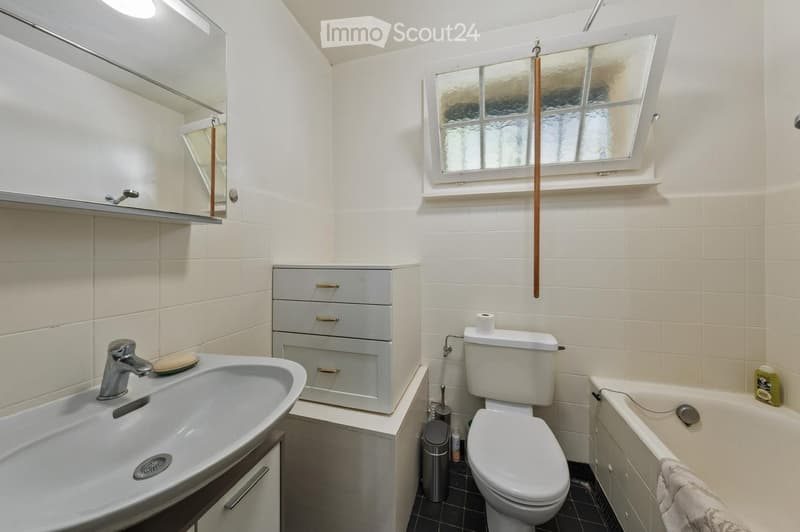 Modern bathroom with white tiles, a sink with a chrome faucet, a toilet, a bathtub, a mirrored cabinet, and a window with frosted glass. A towel hangs on the side of the bathtub.