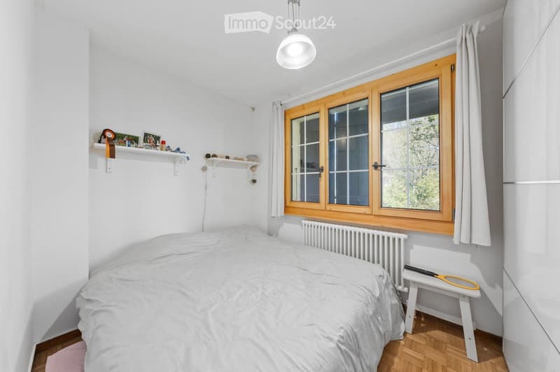 Cozy bedroom with a white bed, wooden floor, large window with wooden frame, shelves with decorative items, and a small stool with a tennis racket on it.