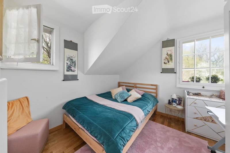 Cozy attic bedroom with wooden bed, teal duvet, nightstand, small round table, and white dresser.