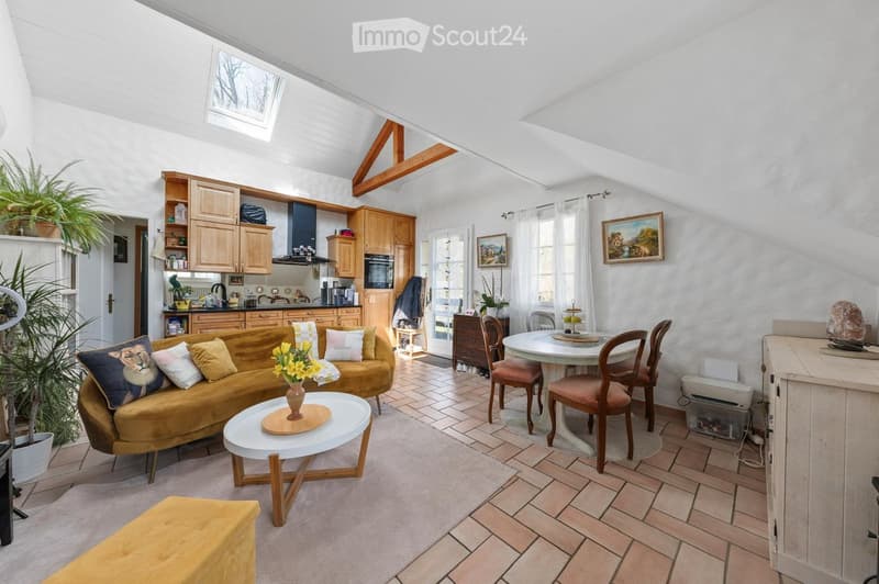 Bright living and dining area with exposed wooden beams, white walls, and tiled flooring. Features a mustard-colored sofa, white coffee table, dining table with chairs, and a small kitchen area with wooden cabinets and stainless steel appliances.