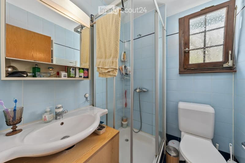 Bathroom with light blue tiles, a white sink with a wooden countertop, a mirror cabinet, a shower area with a glass door, and a toilet. A towel hangs on the shower door, and a toothbrush holder is on the sink.