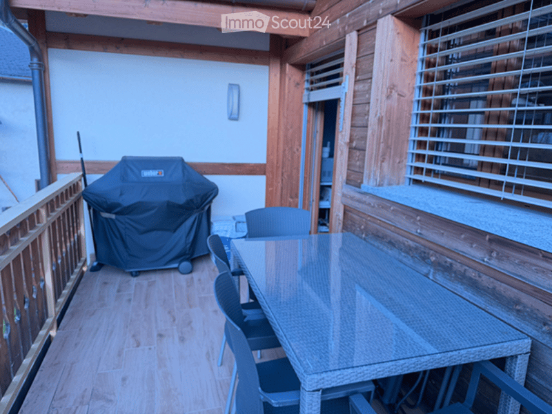Wooden balcony with a glass table, chairs, and a covered grill, featuring wooden railings and a light fixture on the wall.