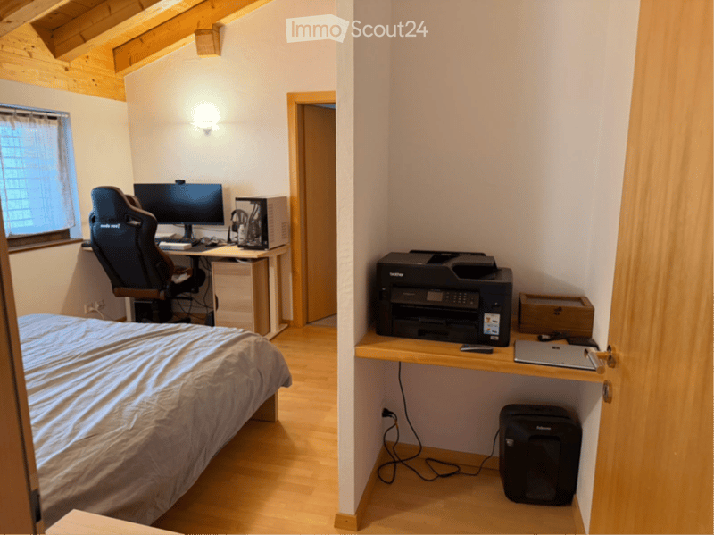 Cozy attic bedroom with wooden beams, a bed, desk with a chair, and a printer on a shelf.