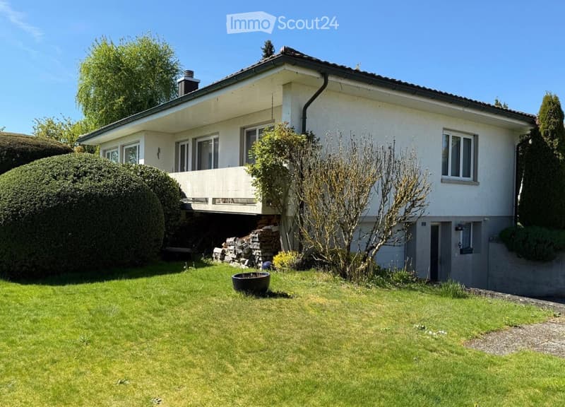 Two-story house with white exterior, multiple windows, and a balcony. The house is surrounded by a well-maintained lawn, shrubs, and trees.