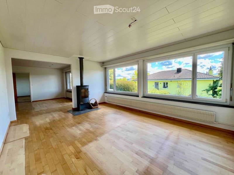 Empty room with wooden flooring, large windows, and a fireplace. The space is well-lit with natural light and has a neutral color scheme.