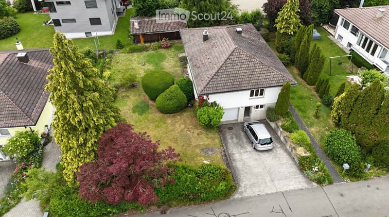 Aerial view of a residential garden area with a house, garage, parked car, and surrounding greenery.