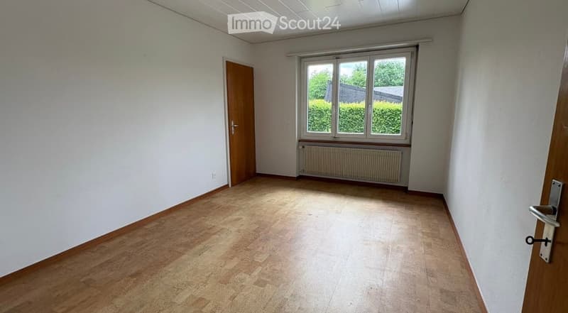 Empty room with light wooden flooring, white walls, a window with white frame, and a wooden door.