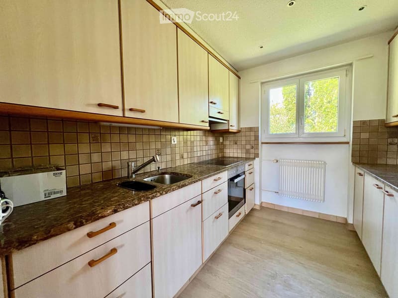 Modern kitchen with white cabinets, granite countertop, sink, faucet, and built-in oven. Light-colored tiles on lower walls, window with view of greenery.