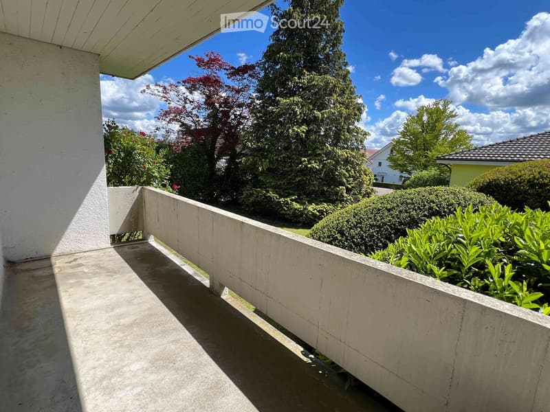 Balcony with concrete railing, overlooking a garden with trees and bushes under a clear blue sky.