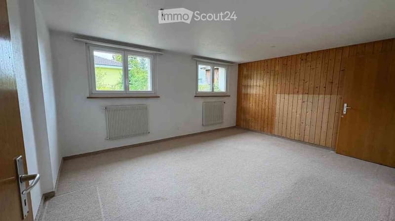 Empty room with light-colored walls, wooden paneling on one side, two windows, radiators, and a tiled floor.