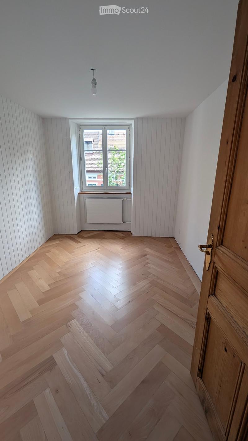 Empty room with wooden herringbone flooring, white paneled walls, a single window with a radiator below, and a hanging light fixture.